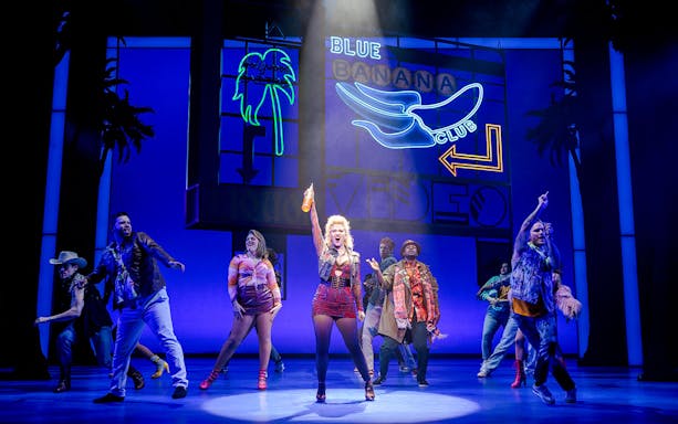 Cast performing a lively scene from Pretty Woman The Musical with colorful neon signs.