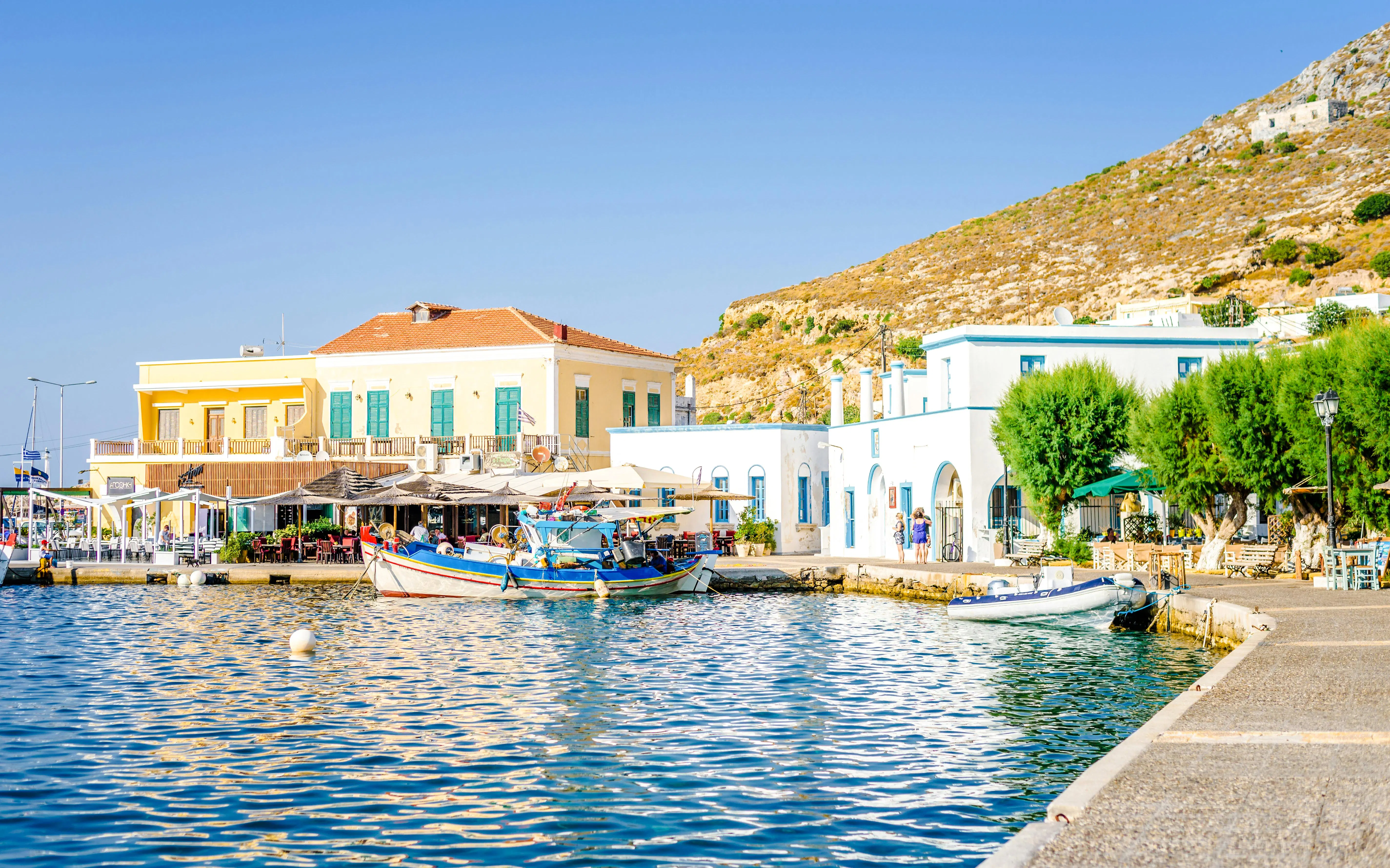 Boats docked at Agia Marina Beach with colorful buildings and hillside in the background.