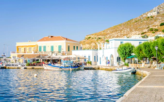 Boats docked at Agia Marina Beach with colorful buildings and hillside in the background.