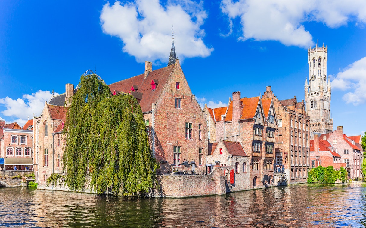 Historic buildings along a canal in Bruges with the Belfry tower in the background.