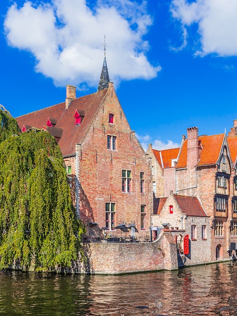 Historic buildings along a canal in Bruges with the Belfry tower in the background.
