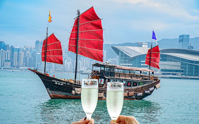 AquaLuna boat with red sails in Hong Kong Harbor, skyline in background, two champagne glasses in foreground.