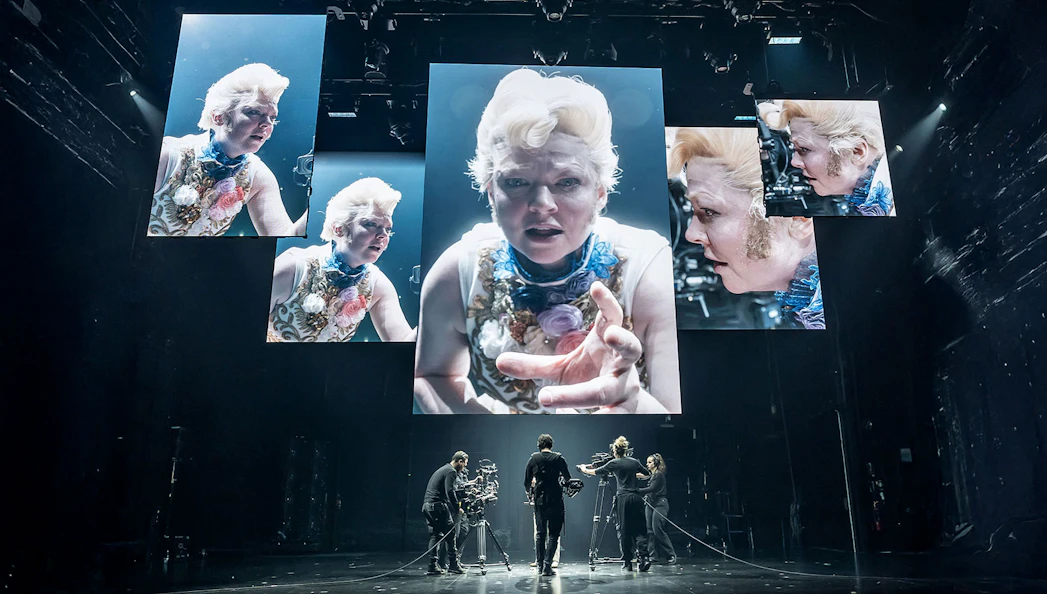 Stage production of "The Picture of Dorian Gray" with actors and large screens displaying close-ups.
