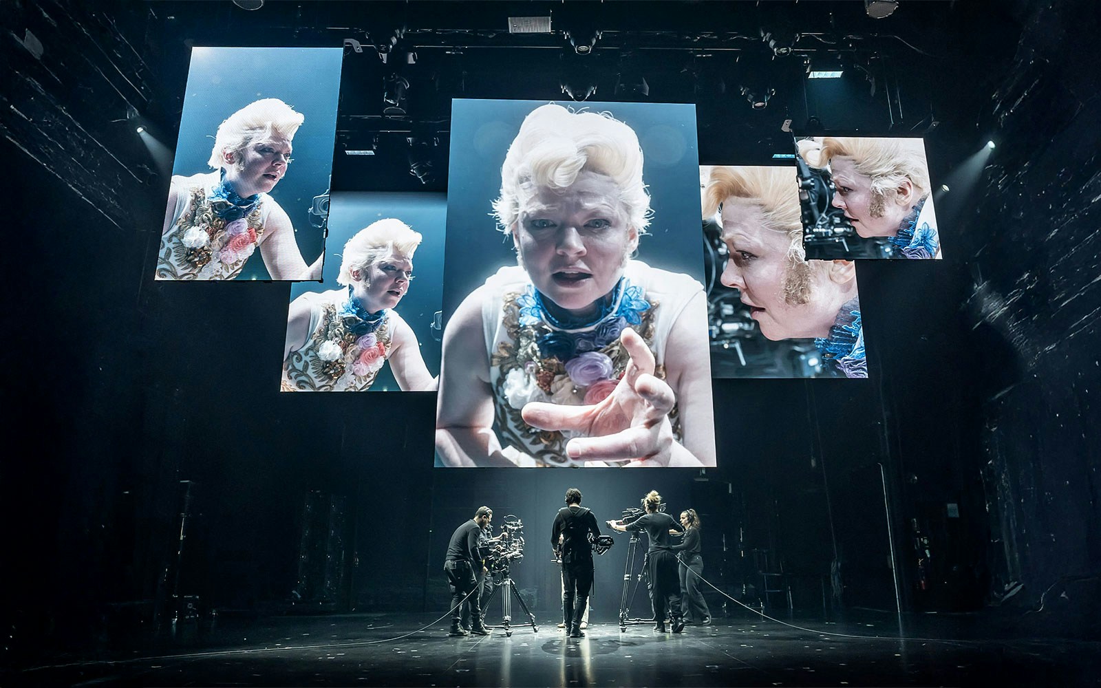 Stage production of "The Picture of Dorian Gray" with actors and large screens displaying close-ups.