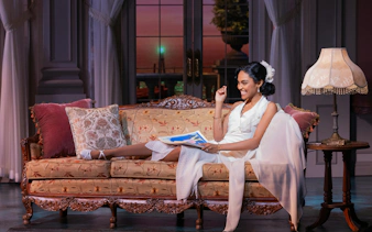 Actor in white dress on vintage sofa, scene from The Great Gatsby musical on Broadway.