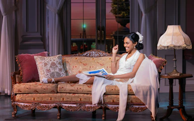 Actor in white dress on vintage sofa, scene from The Great Gatsby musical on Broadway.