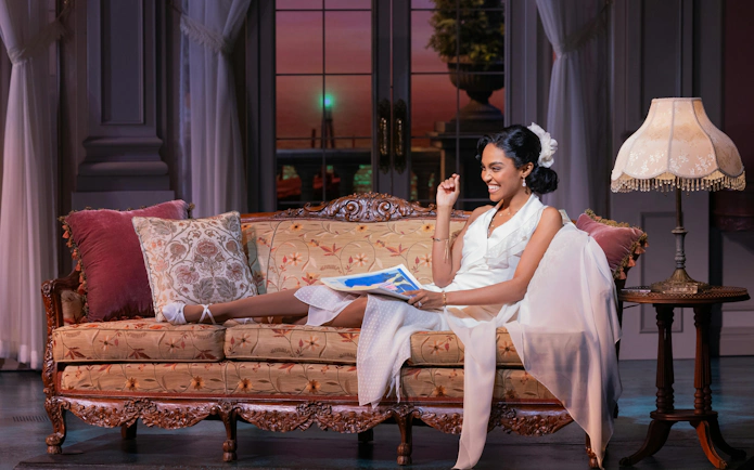 Actor in white dress on vintage sofa, scene from The Great Gatsby musical on Broadway.