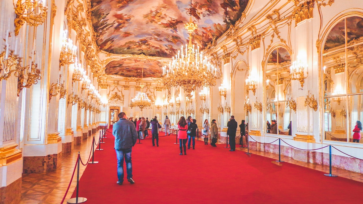Visitors in the grand hall of Schönbrunn Palace, Vienna, with ornate chandeliers and frescoed ceiling.
