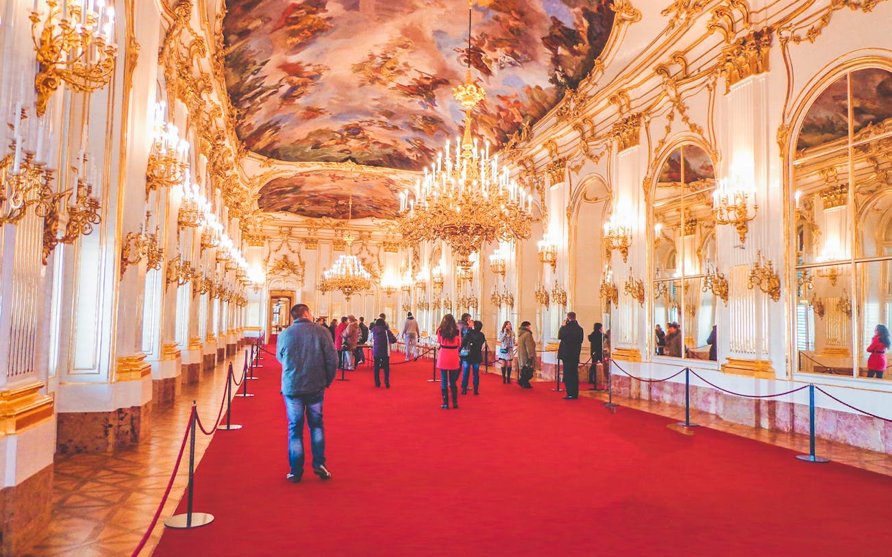 Visitors in the grand hall of Schönbrunn Palace, Vienna, with ornate chandeliers and frescoed ceiling.