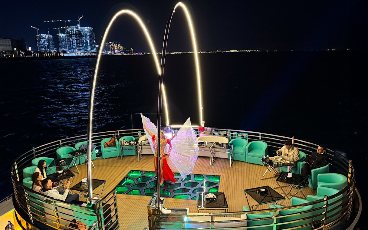 Performer with illuminated wings on a nighttime cruise deck, city lights in the background.