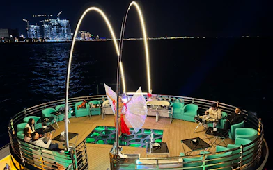Performer with illuminated wings on a nighttime cruise deck, city lights in the background.