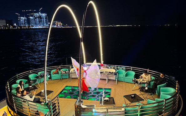 Performer with illuminated wings on a nighttime cruise deck, city lights in the background.