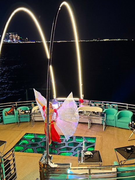 Performer with illuminated wings on a nighttime cruise deck, city lights in the background.