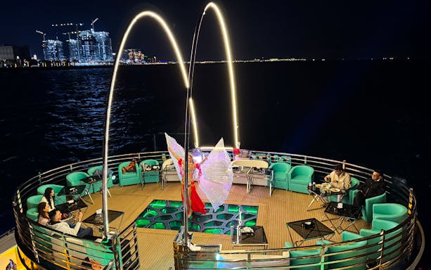 Performer with illuminated wings on a nighttime cruise deck, city lights in the background.