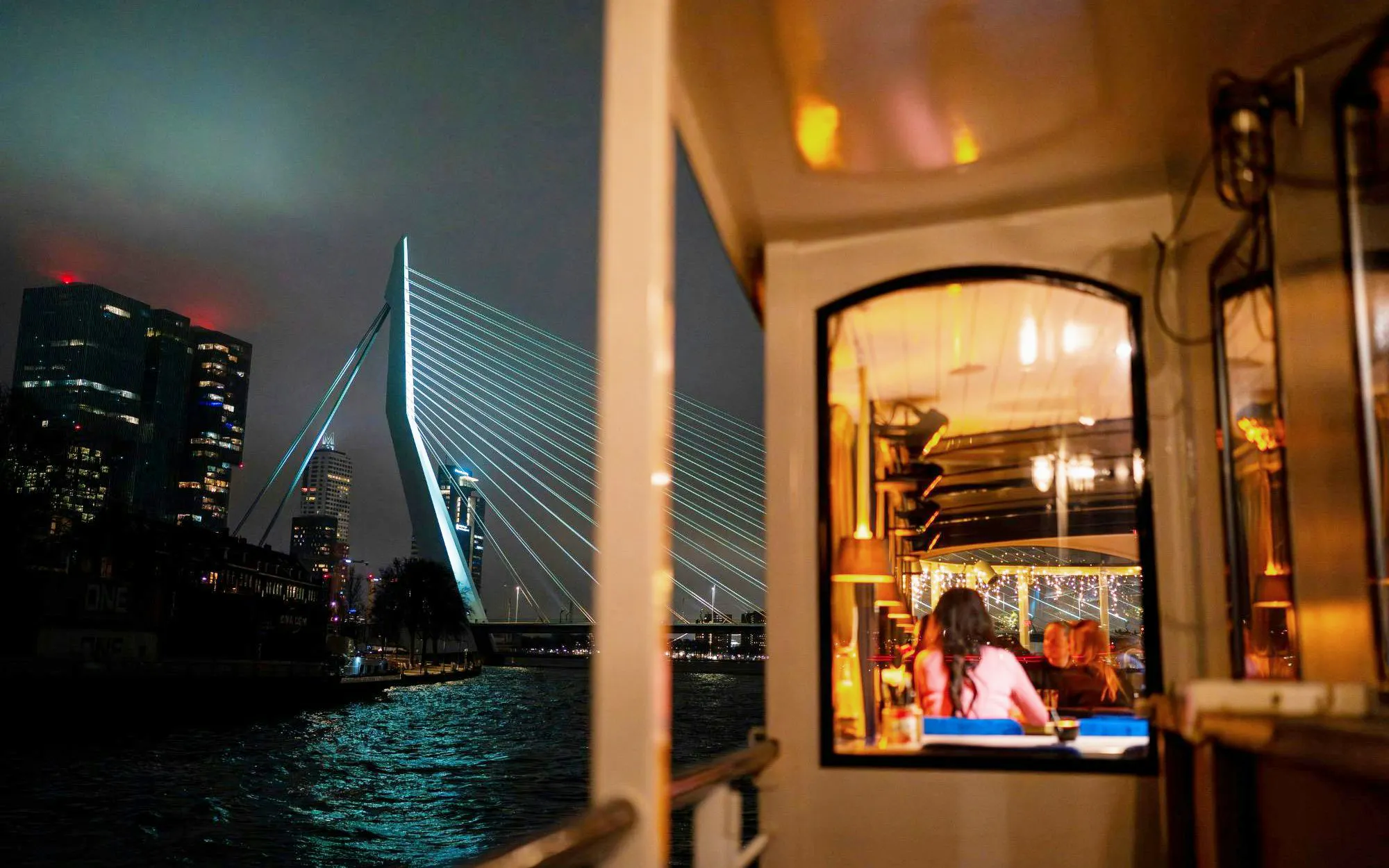 Cruise boat dining with view of Erasmus Bridge in Rotterdam at night.