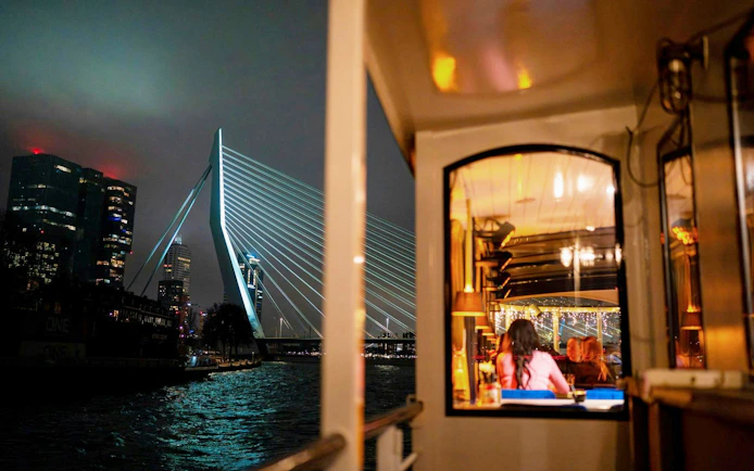 Cruise boat dining with view of Erasmus Bridge in Rotterdam at night.
