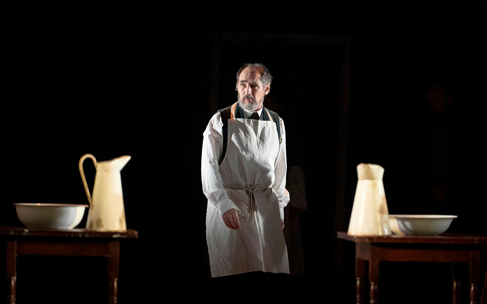 Man in apron standing between tables with washbasins and pitchers, related to Dr Semmelweis.