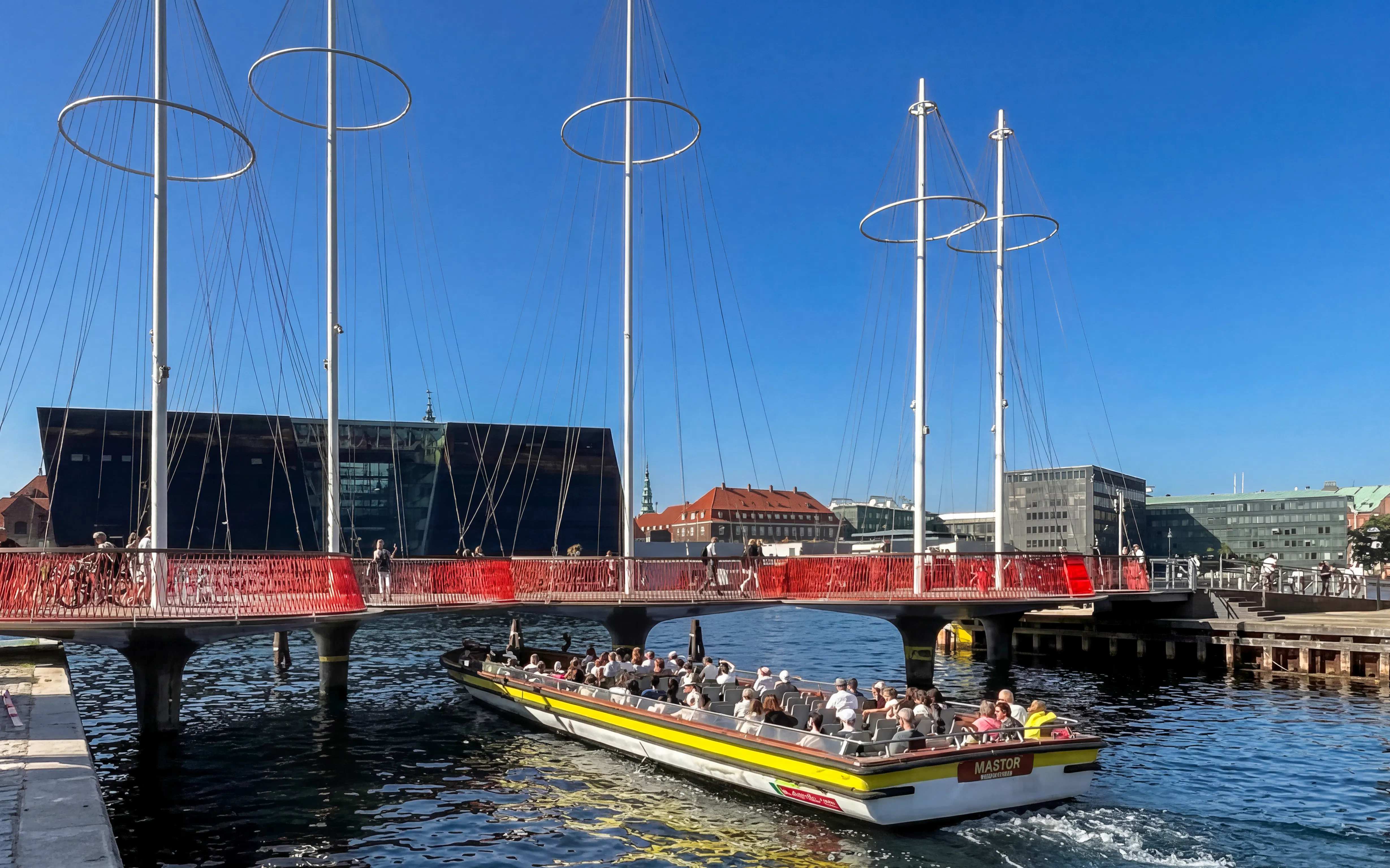 Copenhagen canal cruise boat passing under modern bridge with cityscape in background.