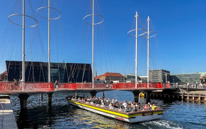 Copenhagen canal cruise boat passing under modern bridge with cityscape in background.