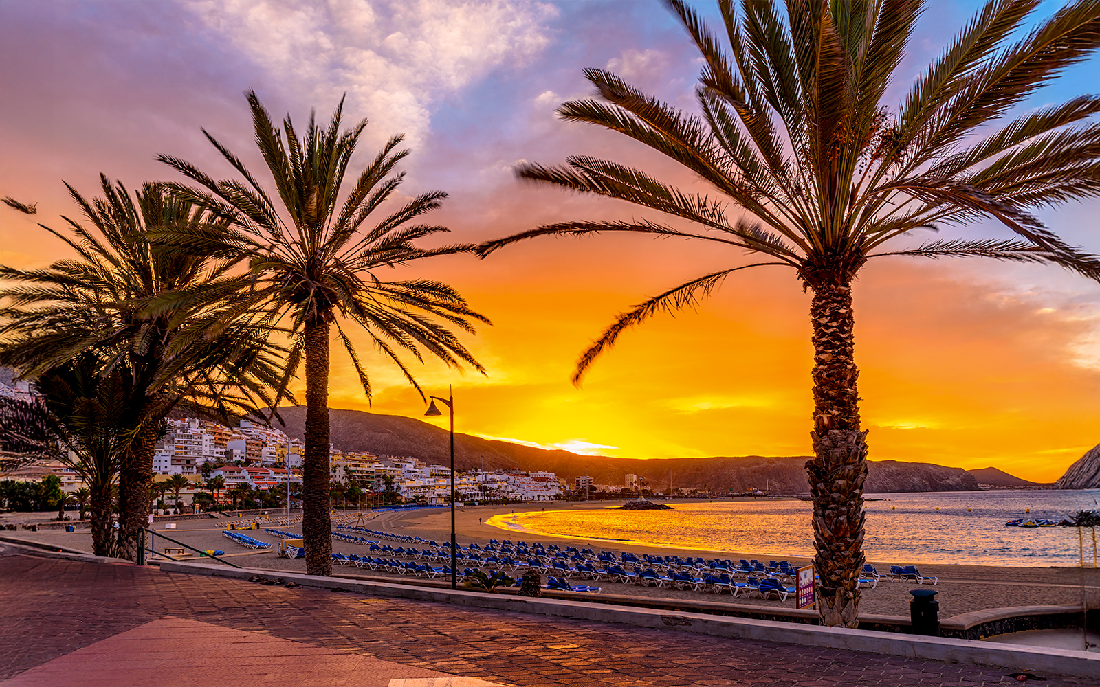 Sunset over Playa de Las Vistas beach with sun loungers and palm trees in Tenerife.