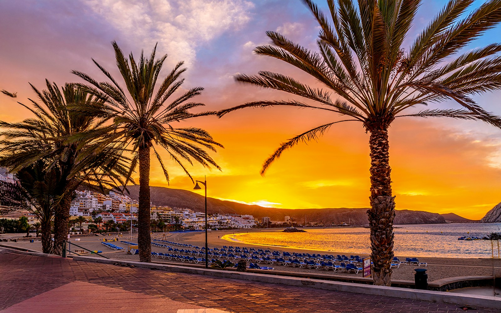 Sunset over Playa de Las Vistas beach with sun loungers and palm trees in Tenerife.