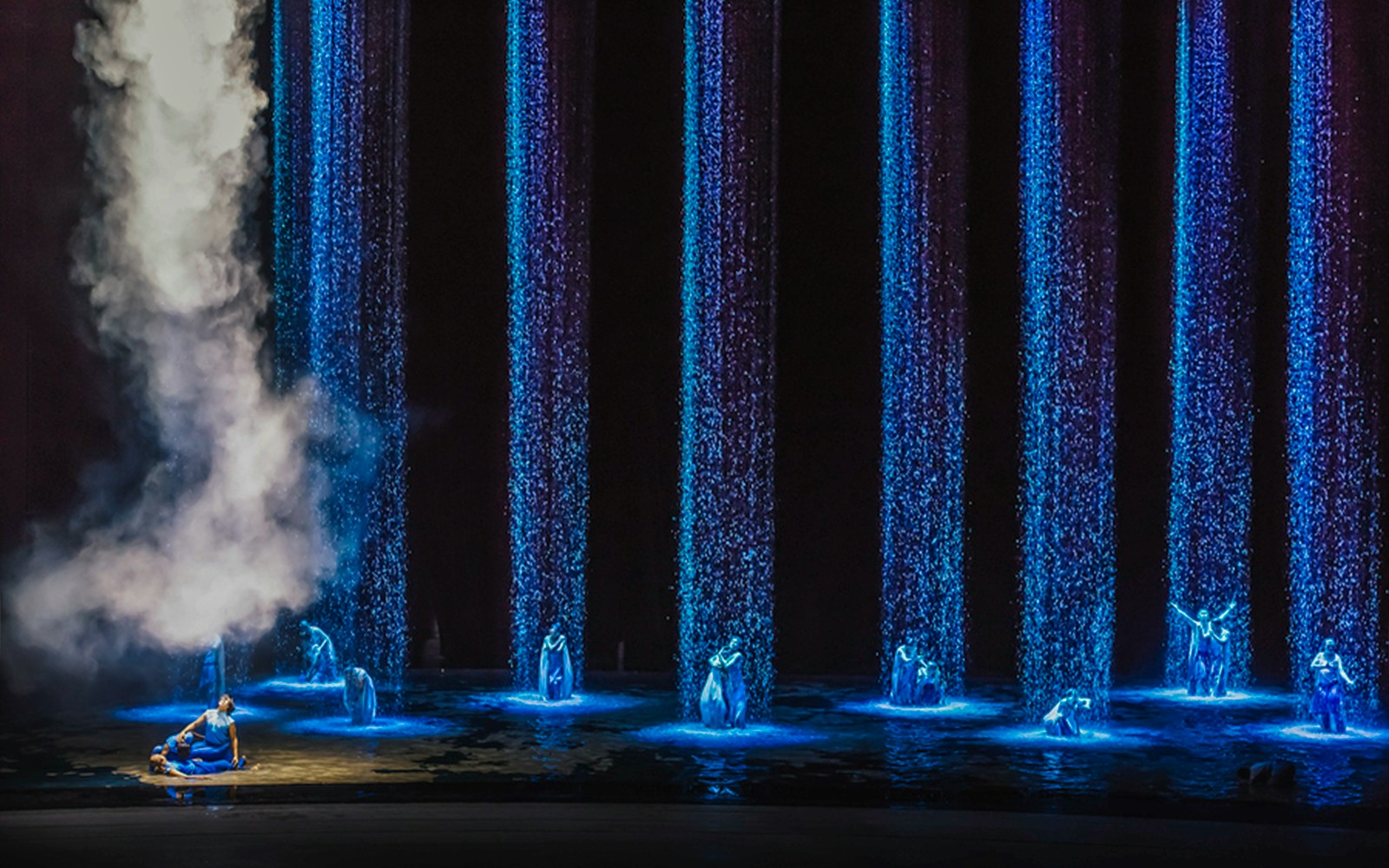 Performers in blue costumes under illuminated water streams and mist.