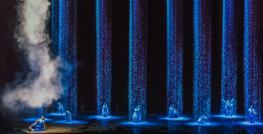 Performers in blue costumes under illuminated water streams and mist.