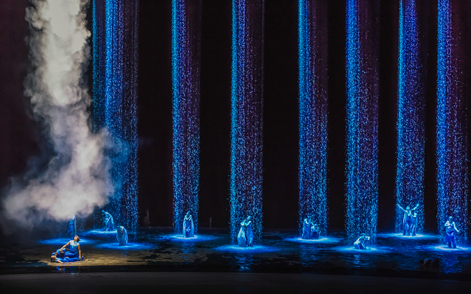Performers in blue costumes under illuminated water streams and mist.