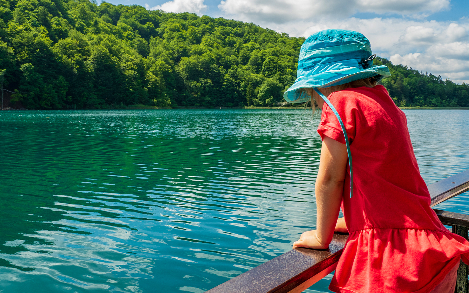 Child in blue hat looking at Plitvice Lakes from a boat, surrounded by lush greenery.