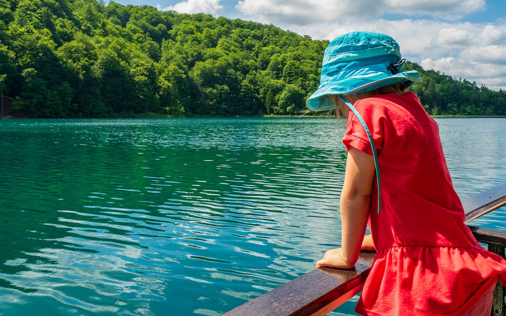 Child in blue hat looking at Plitvice Lakes from a boat, surrounded by lush greenery.
