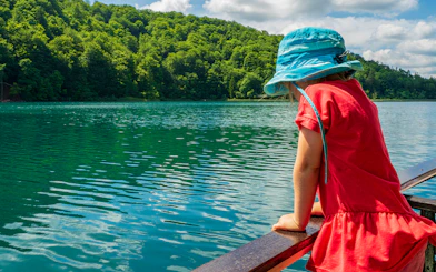 Child in blue hat looking at Plitvice Lakes from a boat, surrounded by lush greenery.