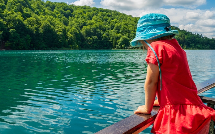 Child in blue hat looking at Plitvice Lakes from a boat, surrounded by lush greenery.