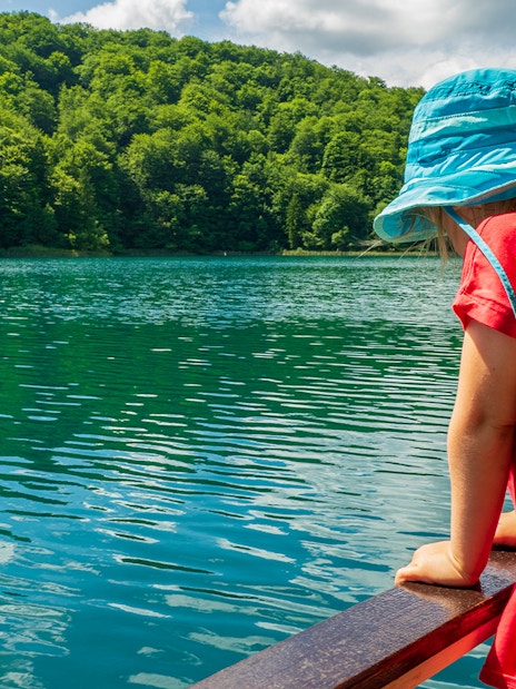 Child in blue hat looking at Plitvice Lakes from a boat, surrounded by lush greenery.