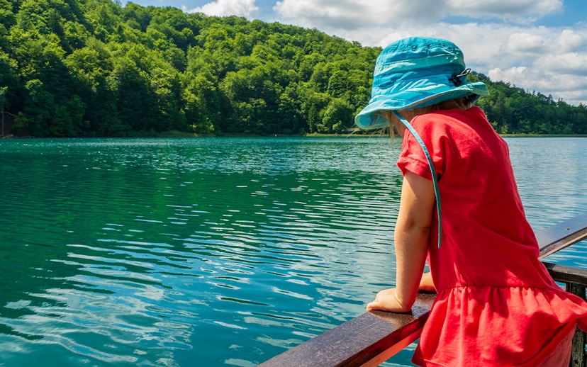Child in blue hat looking at Plitvice Lakes from a boat, surrounded by lush greenery.