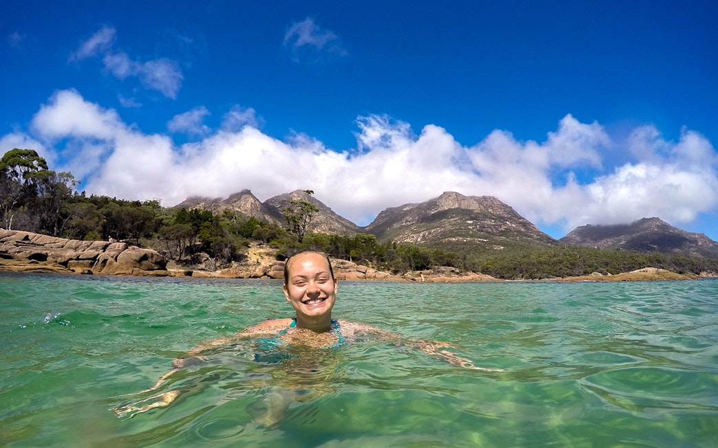 Person swimming in Wineglass Bay with Freycinet National Park mountains in the background.