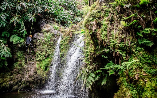 Person rappelling down a waterfall surrounded by lush greenery at Ribeira dos Caldeirões.