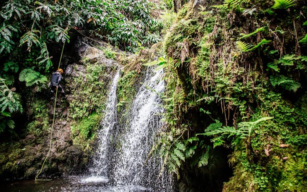 Person rappelling down a waterfall surrounded by lush greenery at Ribeira dos Caldeirões.