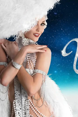 Showgirl in feathered costume with "Vegas! The Show" text on starry background.