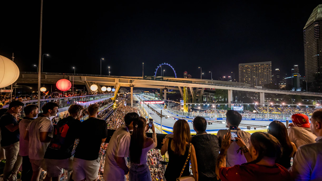 Crowd watching a night race with city skyline and Ferris wheel in the background.