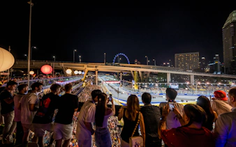 Crowd watching a night race with city skyline and Ferris wheel in the background.