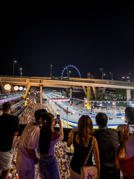 Crowd watching a night race with city skyline and Ferris wheel in the background.