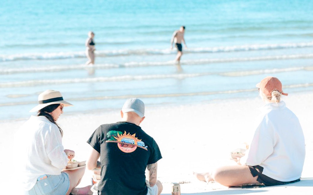 Three people enjoying lunch on Hervey Bay's white sand beach with turquoise ocean.