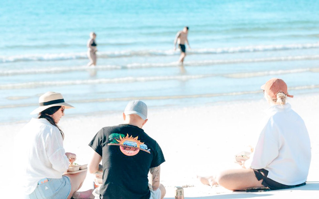 Three people enjoying lunch on Hervey Bay's white sand beach with turquoise ocean.