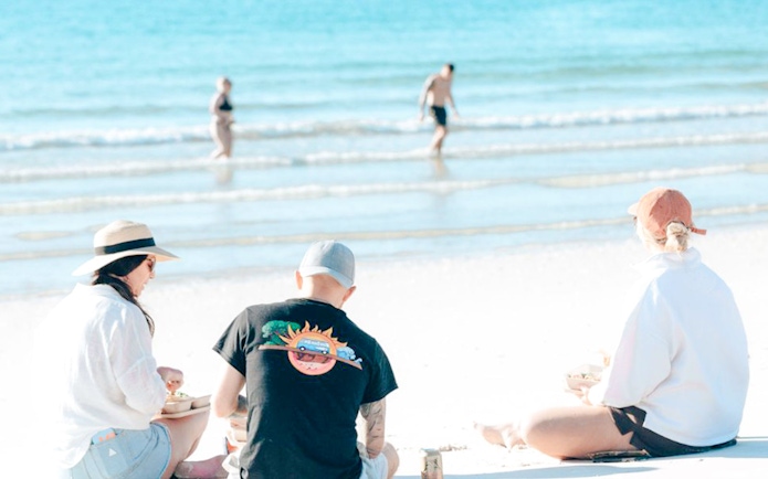 Three people enjoying lunch on Hervey Bay's white sand beach with turquoise ocean.