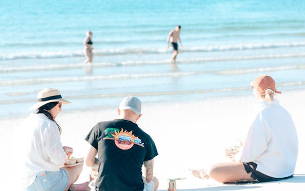 Three people enjoying lunch on Hervey Bay's white sand beach with turquoise ocean.