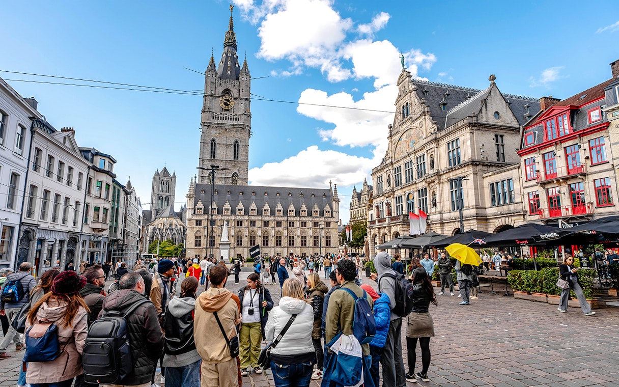 Tourists gather in Ghent's historic center with the Belfry of Ghent in the background.