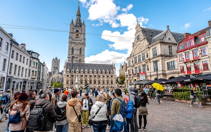 Tourists gather in Ghent's historic center with the Belfry of Ghent in the background.