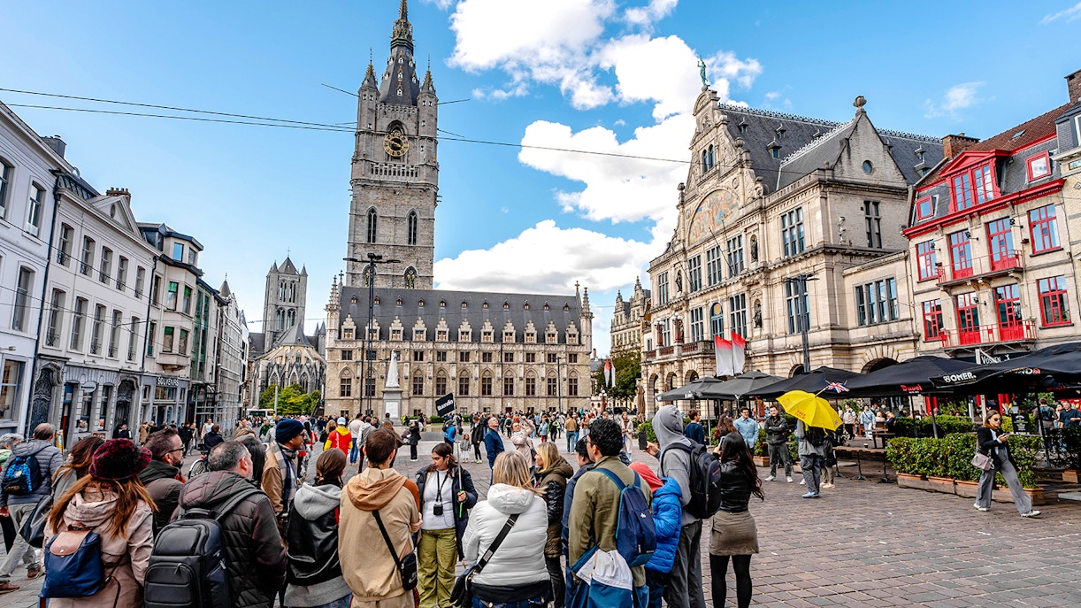 Tourists gather in Ghent's historic center with the Belfry of Ghent in the background.
