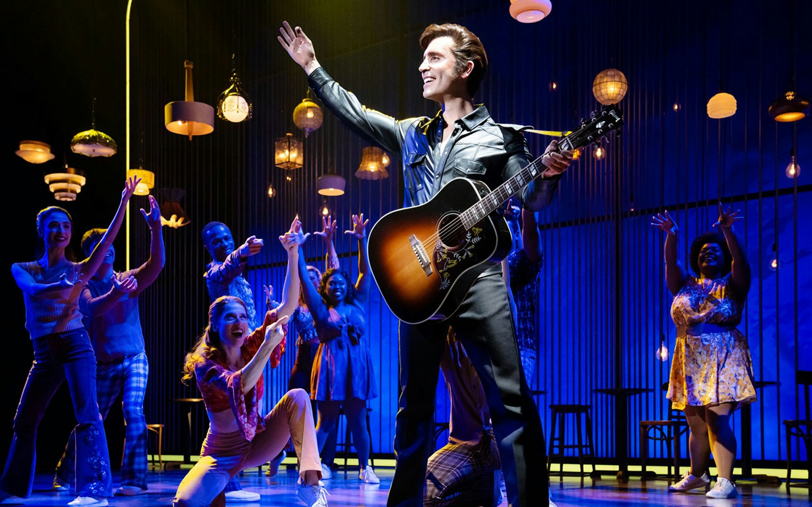 Performer with guitar on stage during "A Beautiful Noise: The Neil Diamond Musical" with ensemble cast.