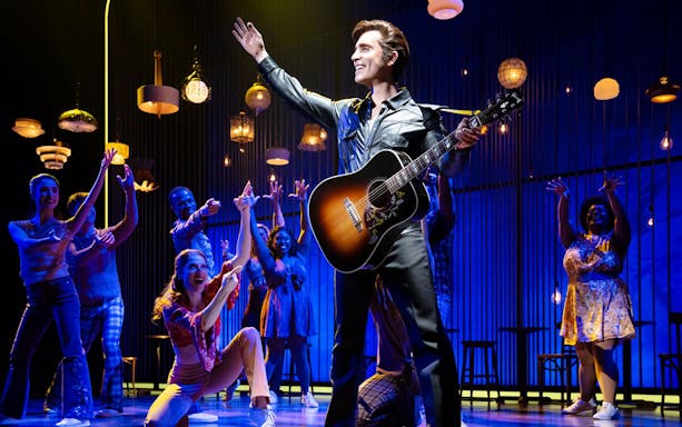 Performer with guitar on stage during "A Beautiful Noise: The Neil Diamond Musical" with ensemble cast.